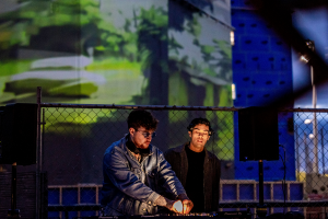 At night, the faces of two electronic musicians are lit from below as they stand against the backdrop of a chain-link fence and, behind that, a campus building façade illuminated by a blurry projection of a green, outdoor scene.