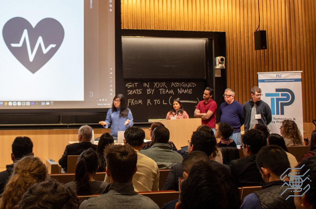 A Policy Hackathon team presents to people in an auditorium, a heart with an EKG line can be seen on the screen